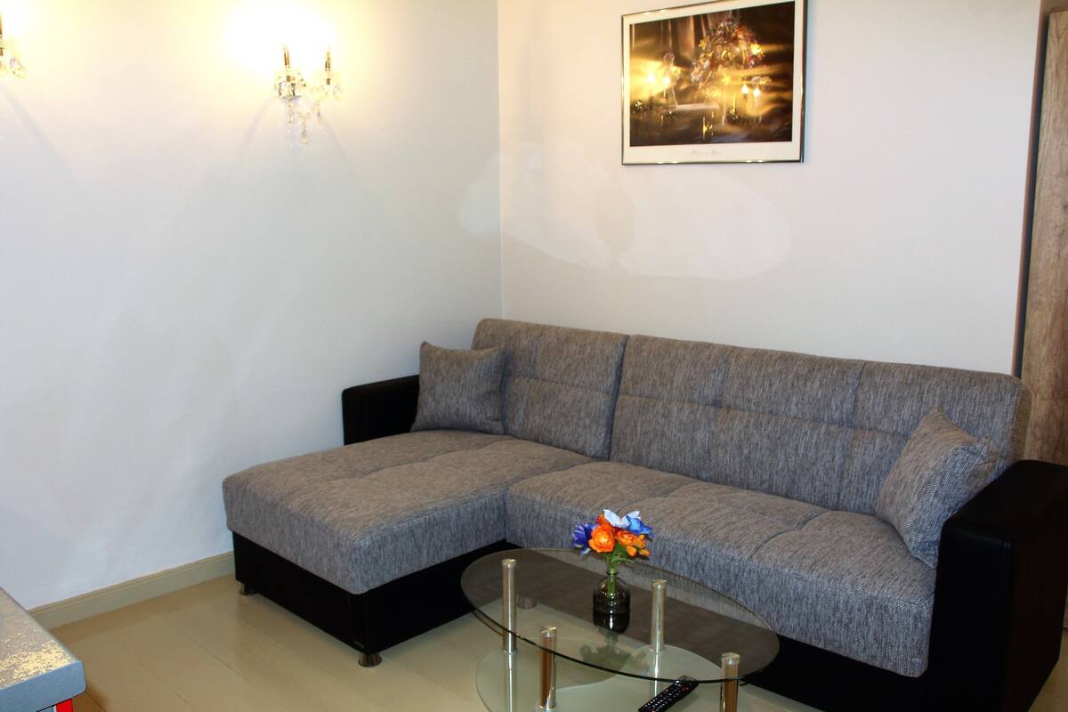 Ein Wohnzimmer mit grauem Sofa, einem Glastisch mit Blumenvase, einem gerahmten Bild an der Wand und einer Deckenleuchte.