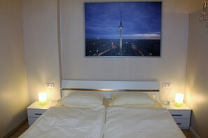 A hotel room with a bed, two bedside tables with lamps, a nightstand, and a framed picture of a tower against a cityscape. - Stars Berlin Apartments Essener Straße (Berlin)