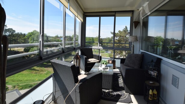 A balcony with wicker furniture, a glass table, and a view of greenery and buildings. - Solferie Holiday Home- Gullveien (Kristiansand)