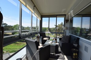 Un balcon agrémenté de meubles en osier, d’une table en verre et offrant une vue sur la verdure et les bâtiments.