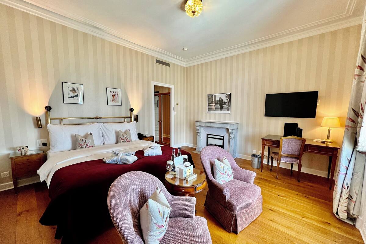 Ein Hotelzimmer mit einem großen Bett, zwei Sesseln, einem Schreibtisch mit Stuhl, einem Kamin, einem Fernseher und einer Tür zu einem Schrank.