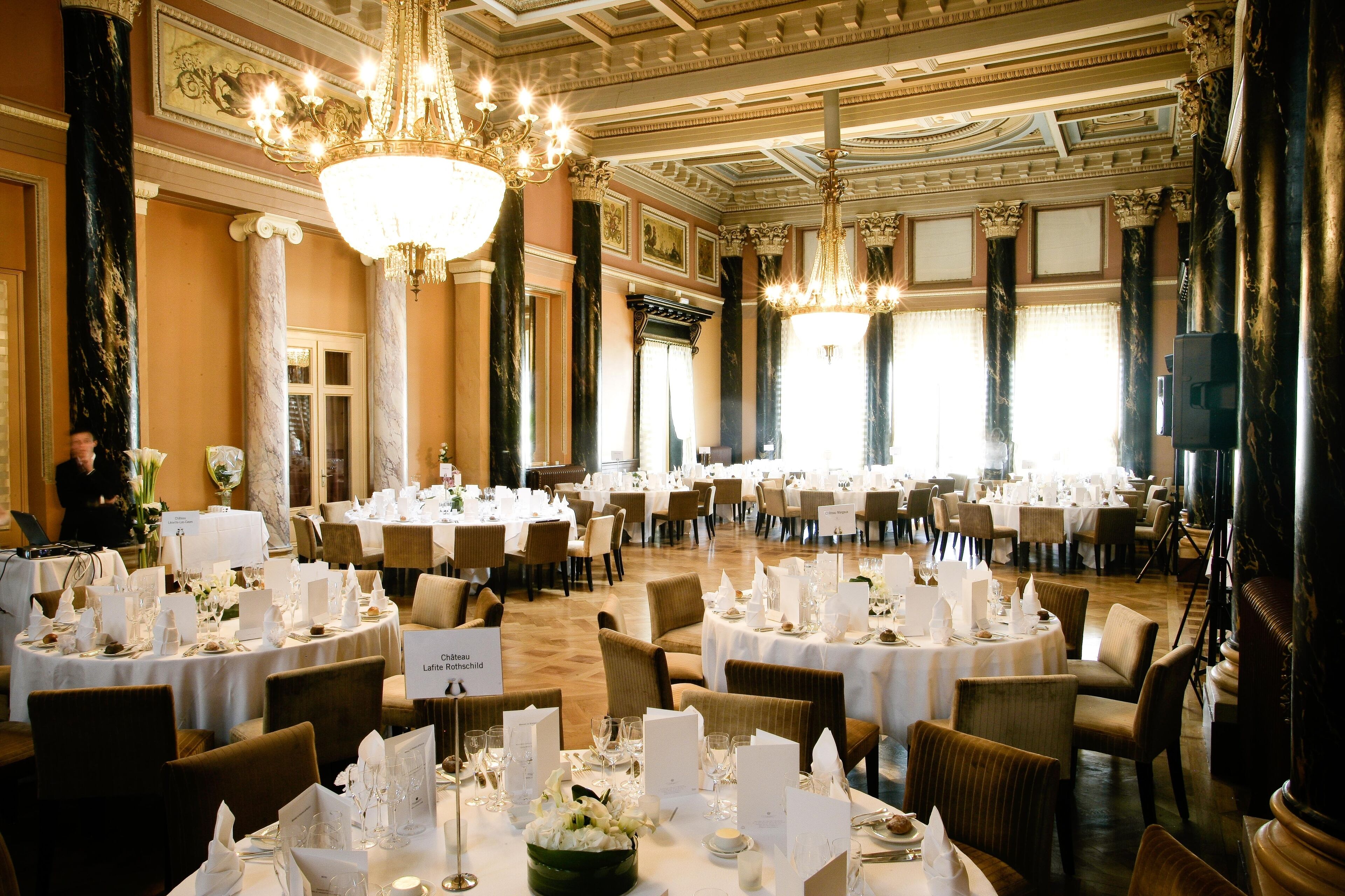 A banquet hall with round tables set for a formal event, featuring chandeliers and classical architecture.