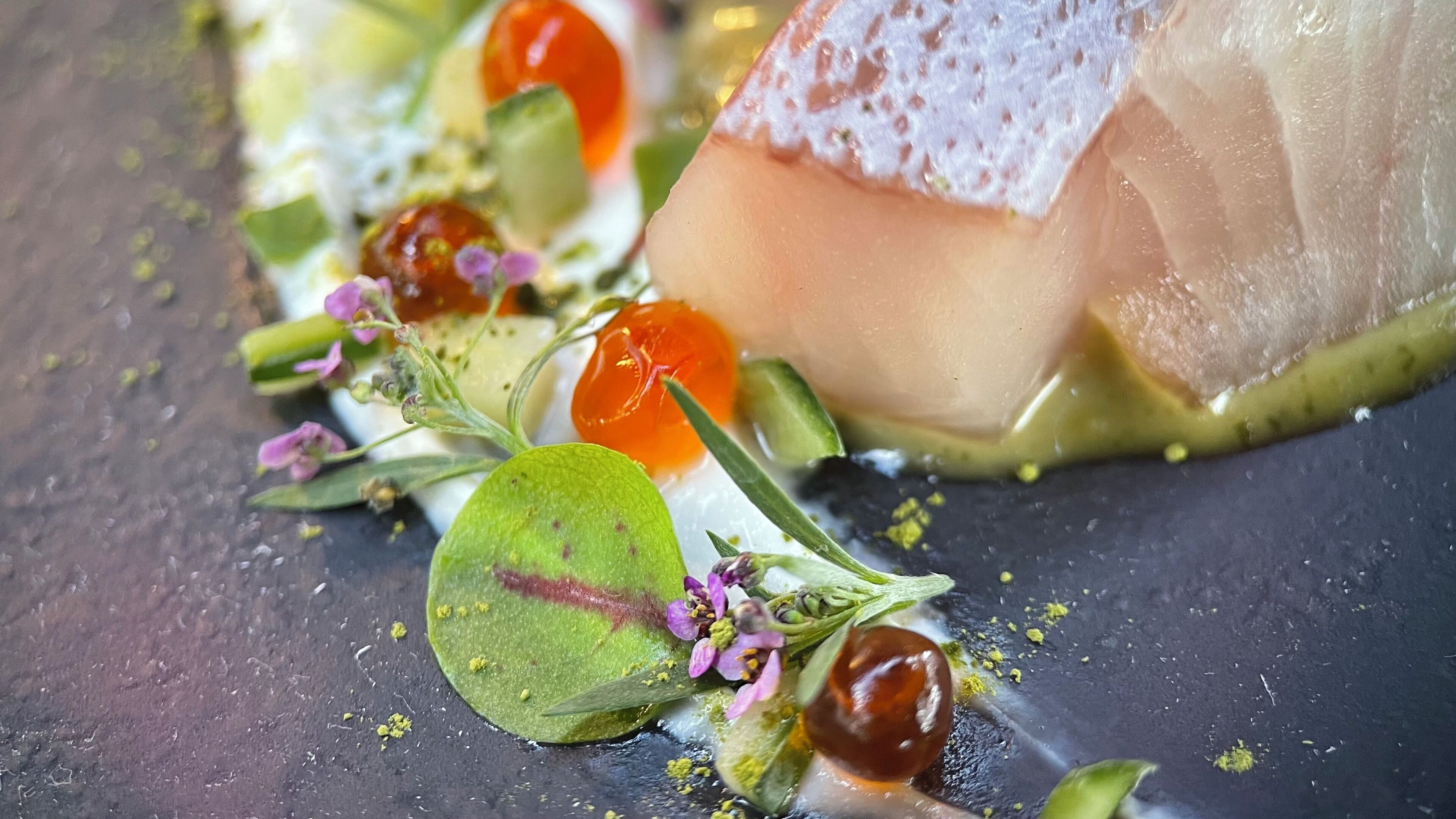 A close-up of a dish with a piece of fish, garnished with green leaves, purple flowers, and a red fruit.