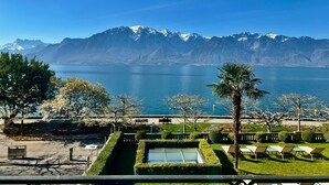 On peut admirer une chaîne de montagnes dont les sommets sont couverts de neige, qui se reflète dans un plan d’eau. Au premier plan, on aperçoit un jardin bien entretenu avec un palmier, ainsi qu’un espace aménagé avec des tables et des chaises.