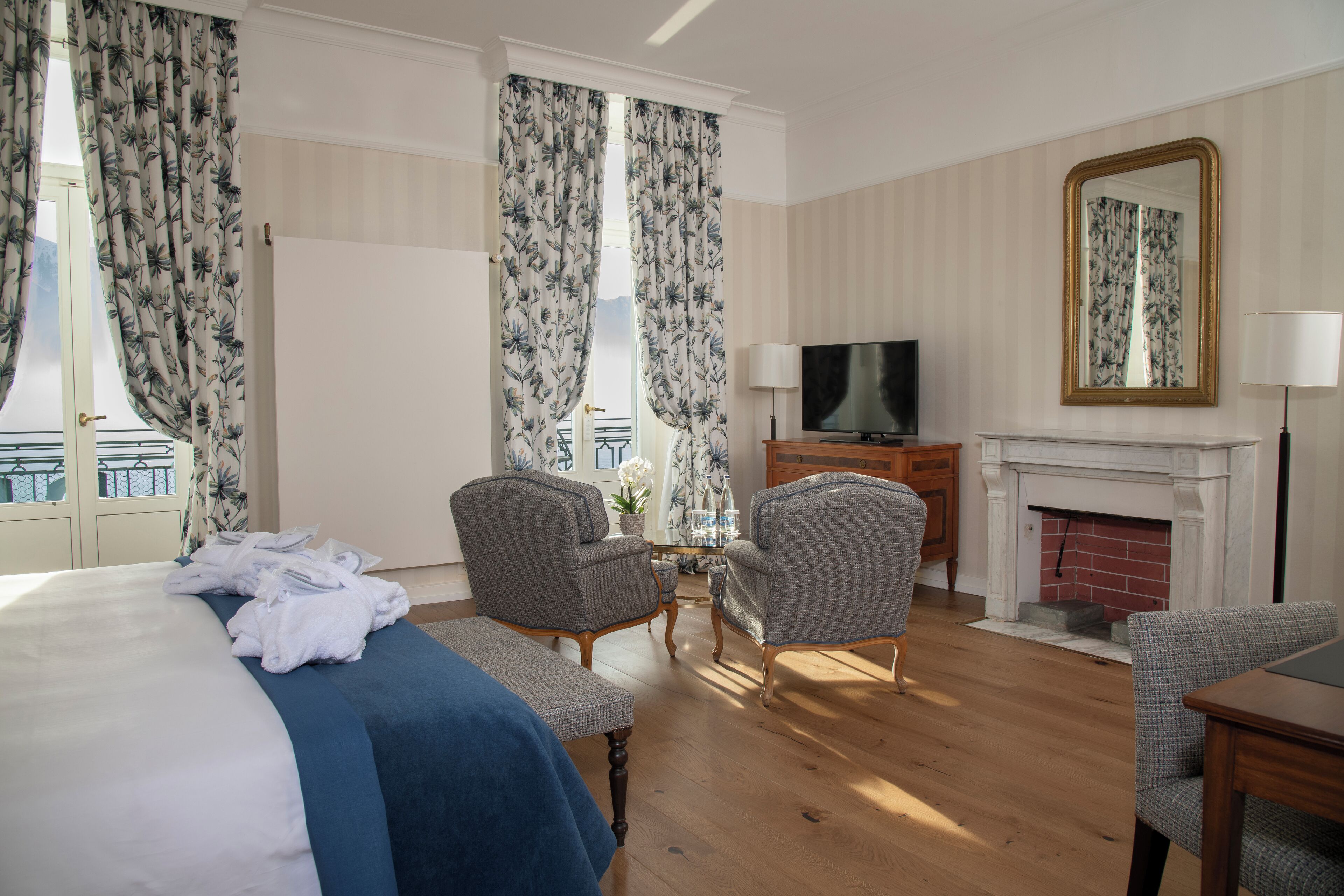 A hotel room with a bed, two armchairs, a television, a fireplace, and a balcony with curtains.