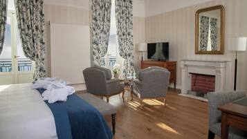 A hotel room with a bed, two armchairs, a television, a fireplace, and a balcony with curtains.