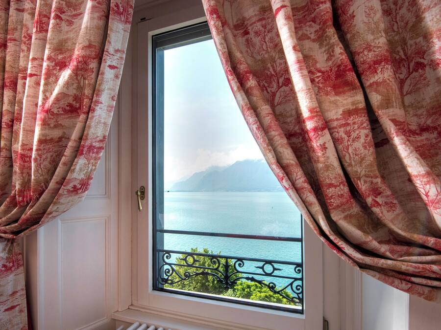 A window with curtains, a view of a body of water, and a balcony railing.