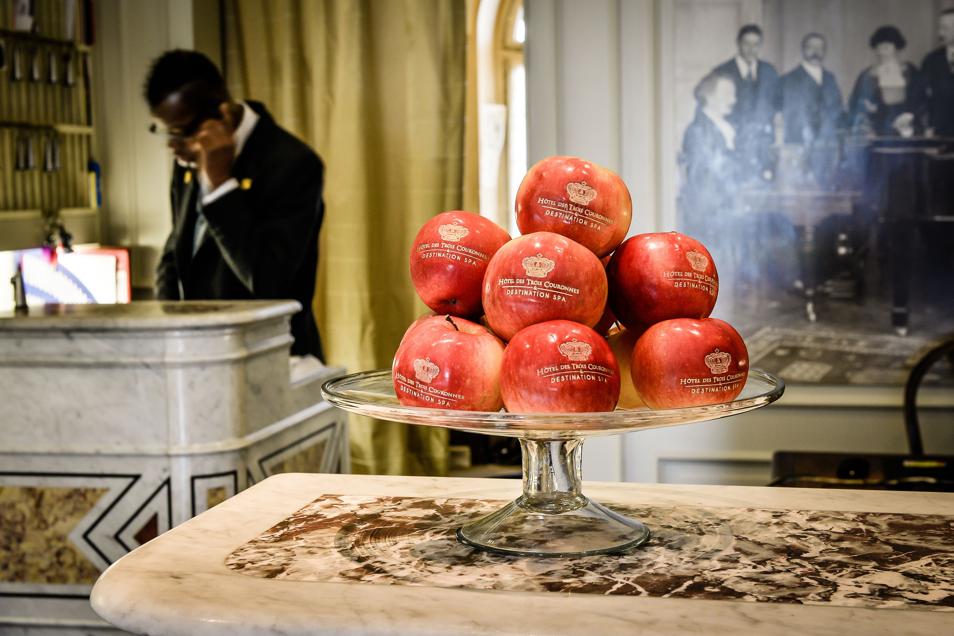 A marble table with a glass stand holding red apples labeled "Hotel du Trou de Cuivre Destination Spa".