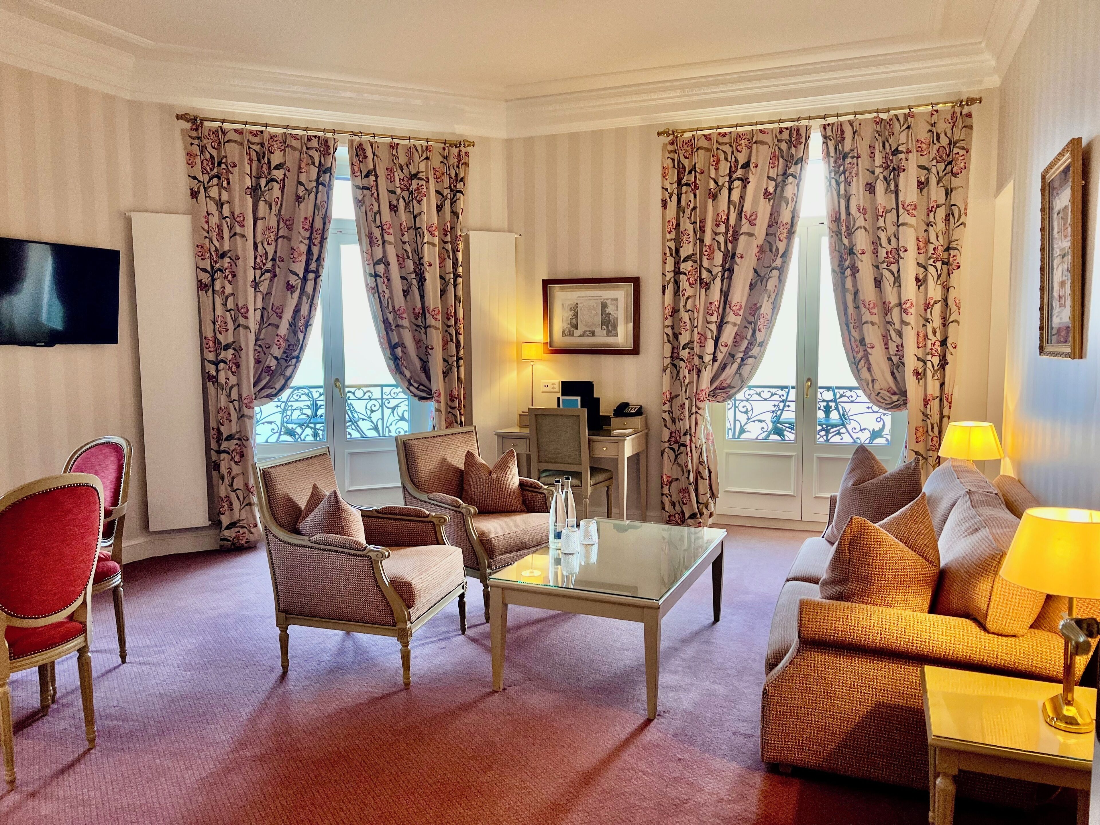 A hotel room with a seating area featuring two armchairs and a sofa, a glass coffee table, a flat-screen TV mounted on the wall, and a desk with a lamp.
