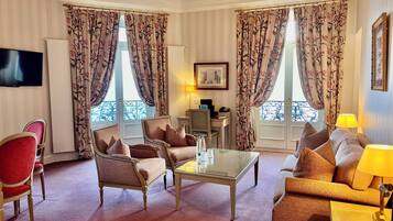 A hotel room with a seating area featuring two armchairs and a sofa, a glass coffee table, a flat-screen TV mounted on the wall, and a desk with a lamp.
