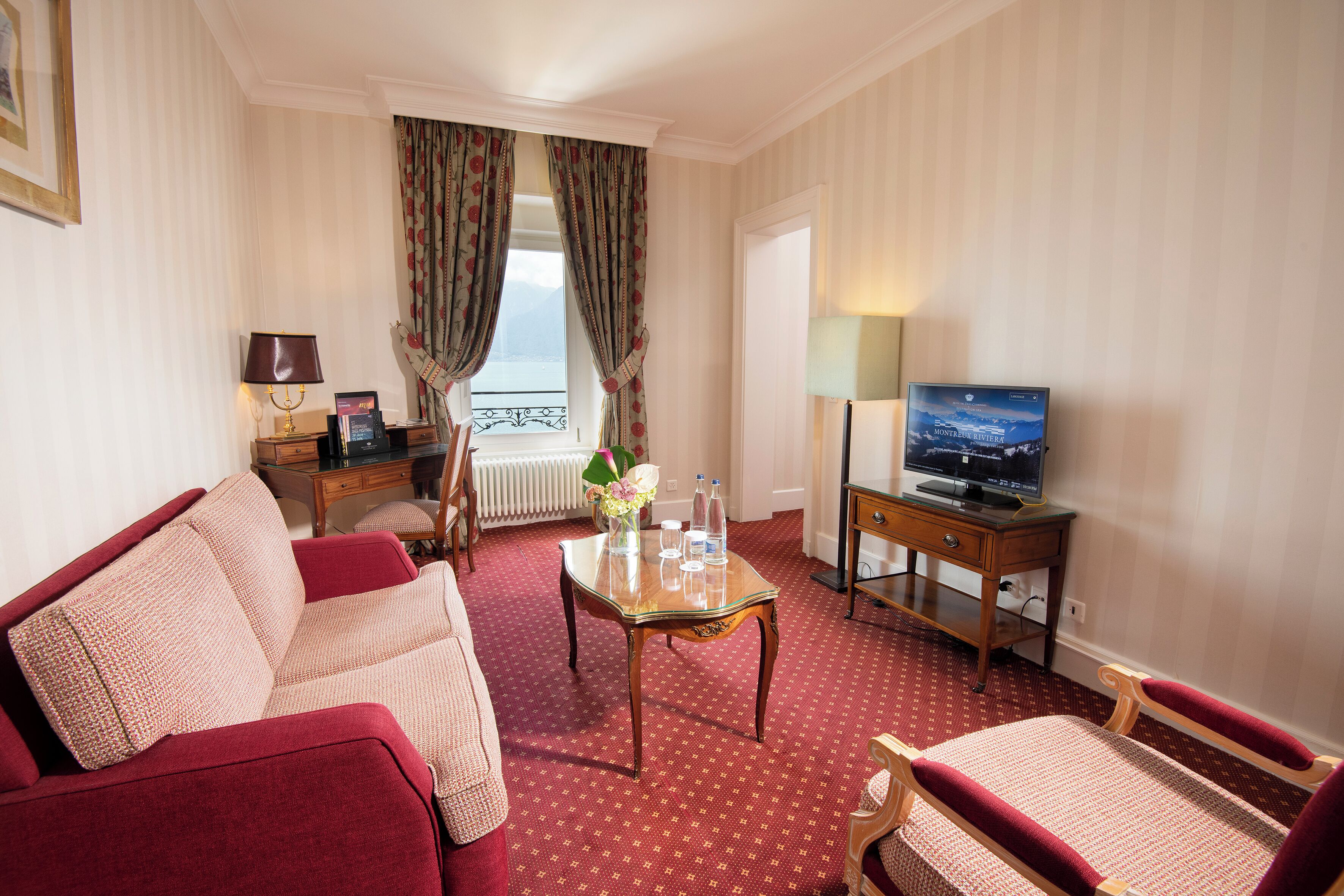 Hotelkamer met een rood tapijt, een bank, een stoel, een kleine tafel met een vaas, een televisie en een raam met gordijnen.