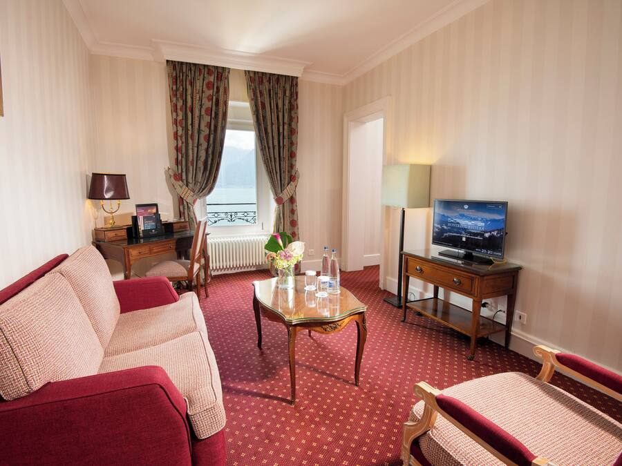 Hotelkamer met een rood tapijt, een bank, een stoel, een kleine tafel met een vaas, een televisie en een raam met gordijnen.