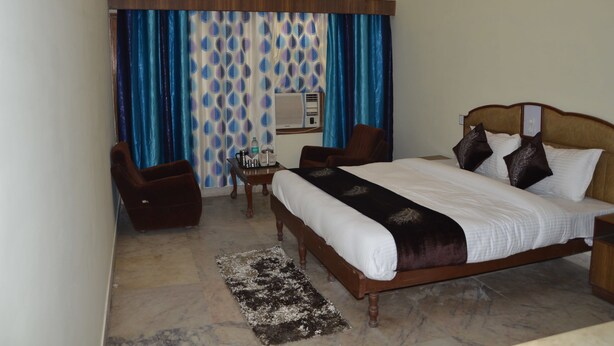 A hotel room with a large bed, a wooden headboard, a small table with a lamp, a chair, and a curtain with a blue and white pattern. - Hotel Vinayakam (Udaipur)