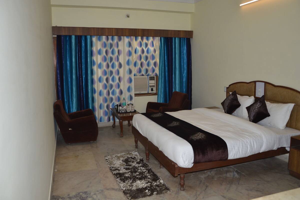 Une chambre d’hôtel avec un grand lit, une tête de lit en bois, une petite table avec une lampe, une chaise et un rideau à motifs bleus et blancs.