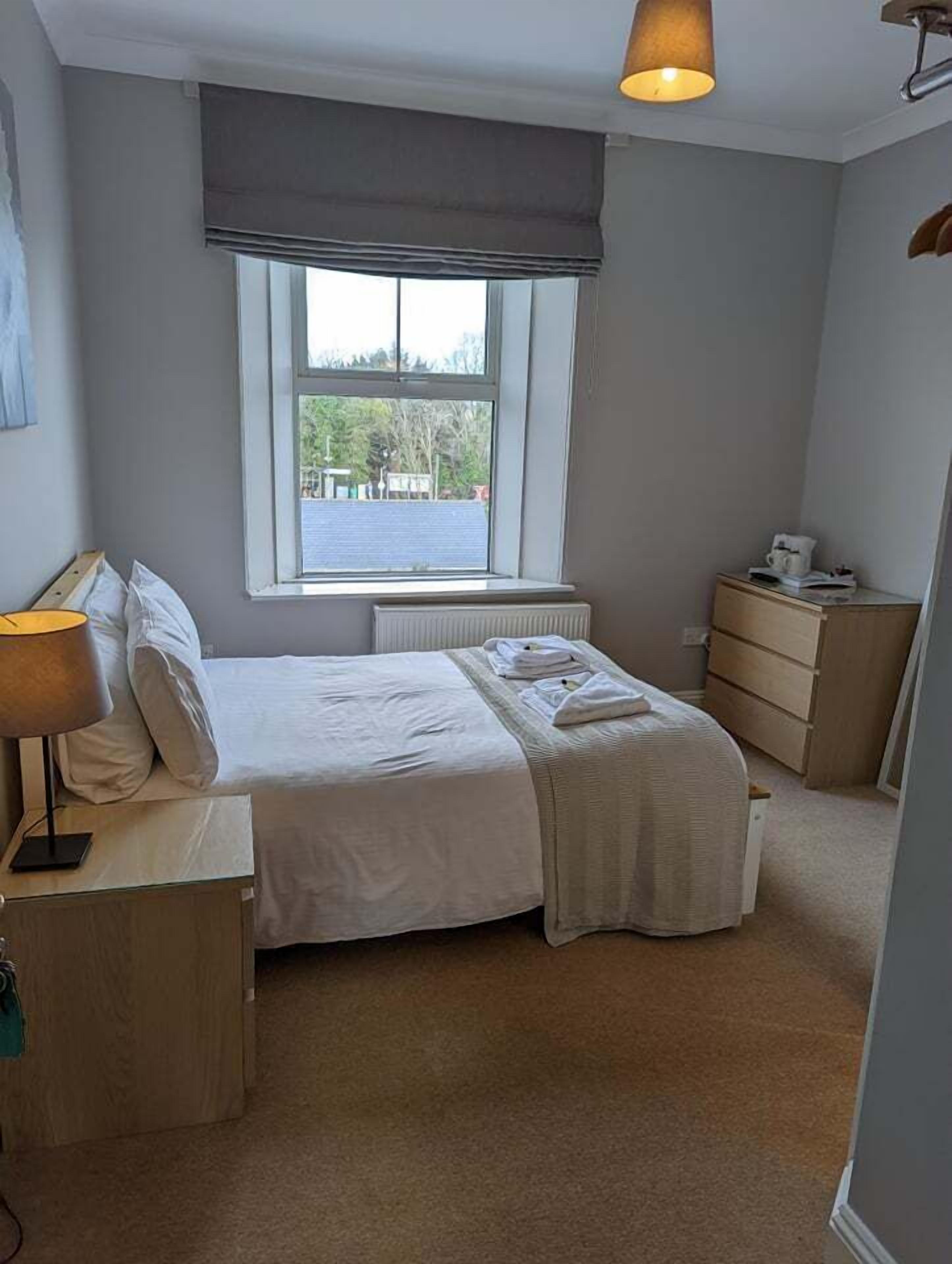 A bedroom with a bed, bedside table, lamp, dresser, and a window with a view of the outside.