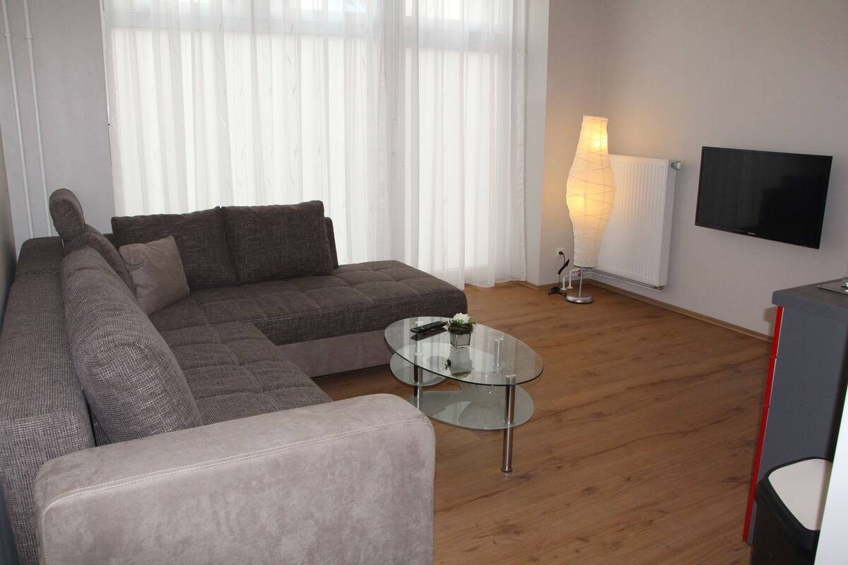 A modern living room with a sectional sofa, a glass coffee table, and a flat-screen TV mounted on the wall.