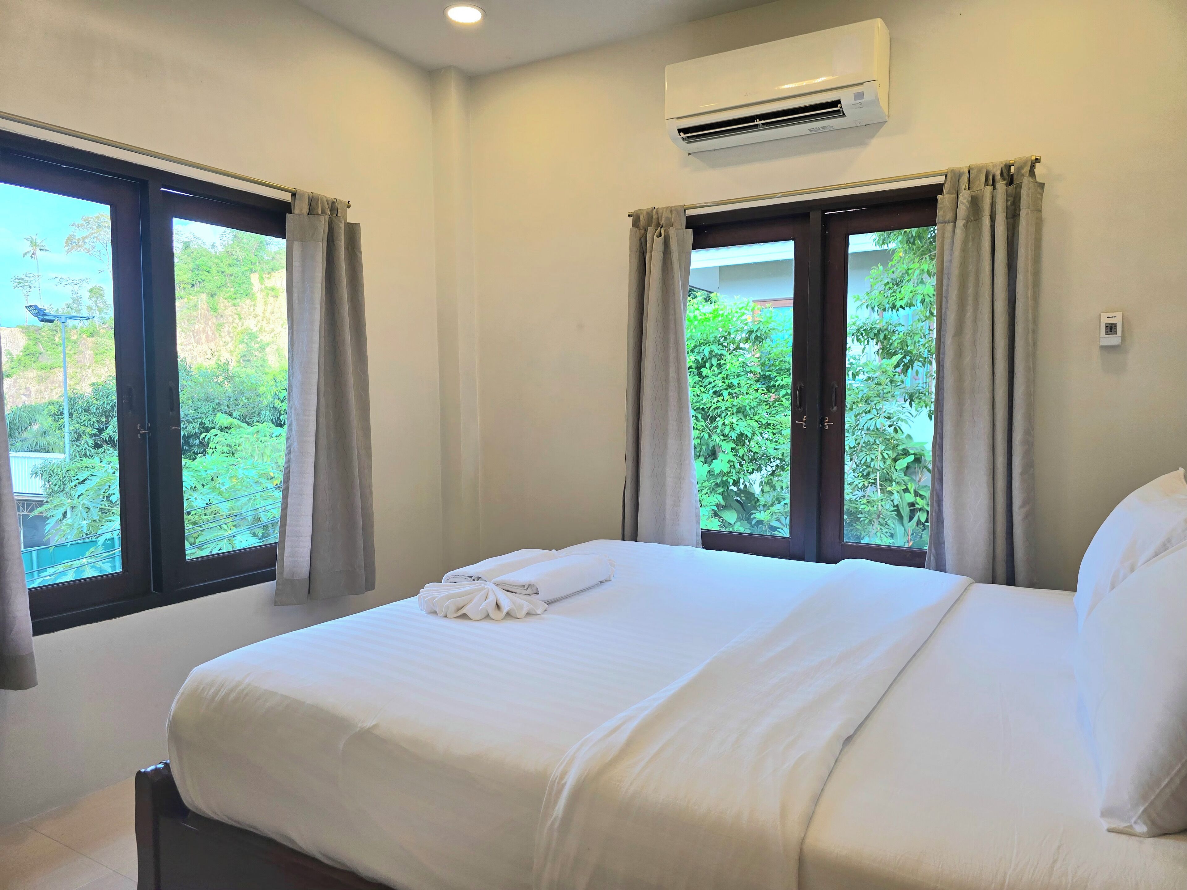 A hotel room with a bed, two windows with curtains, an air conditioning unit, and a view of greenery outside.