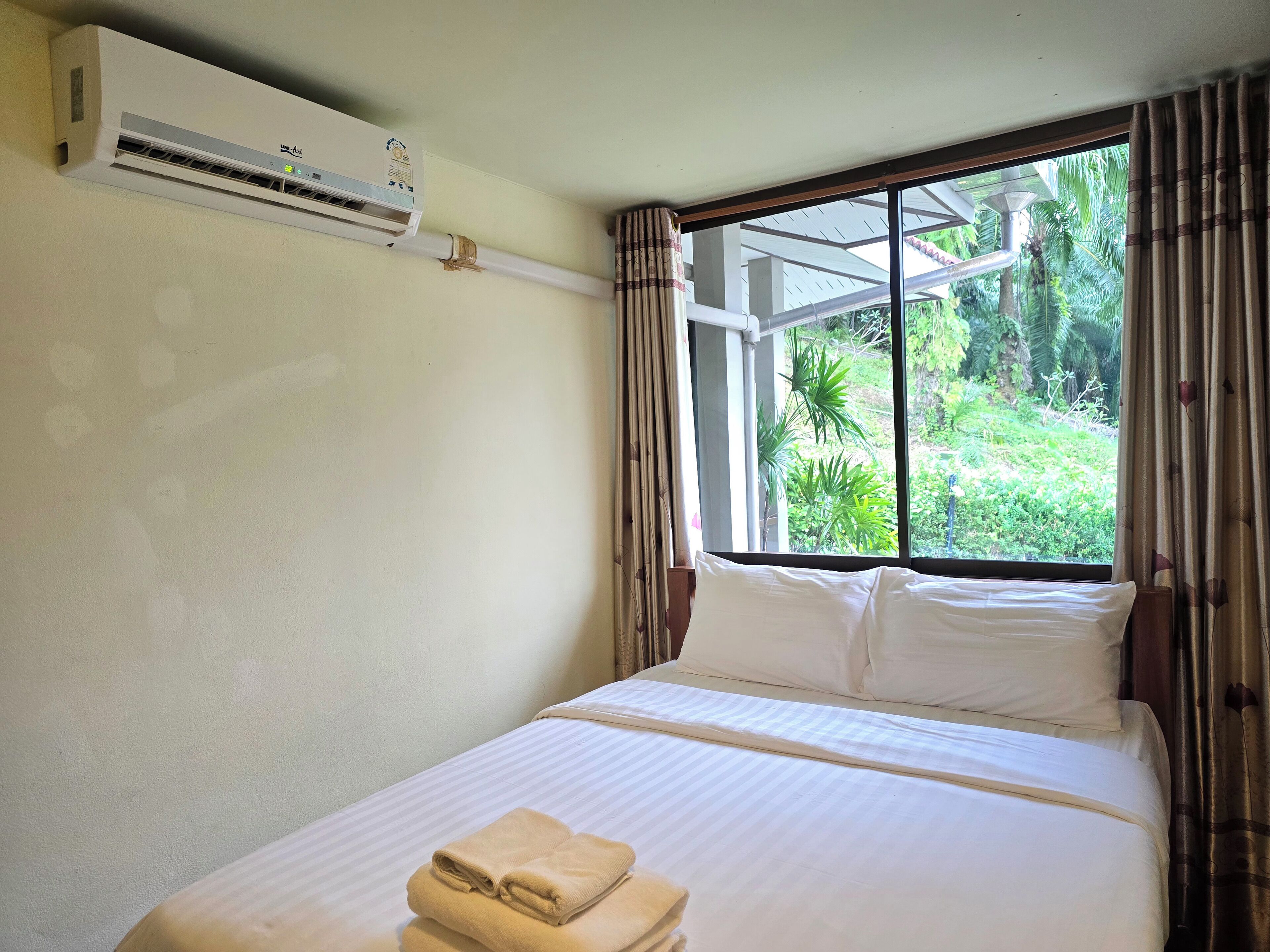 Ein Hotelzimmer mit Bett, einem fenster mit Vorhängen, einer Klimaanlage an der Wand und Blick auf Grünfläche.