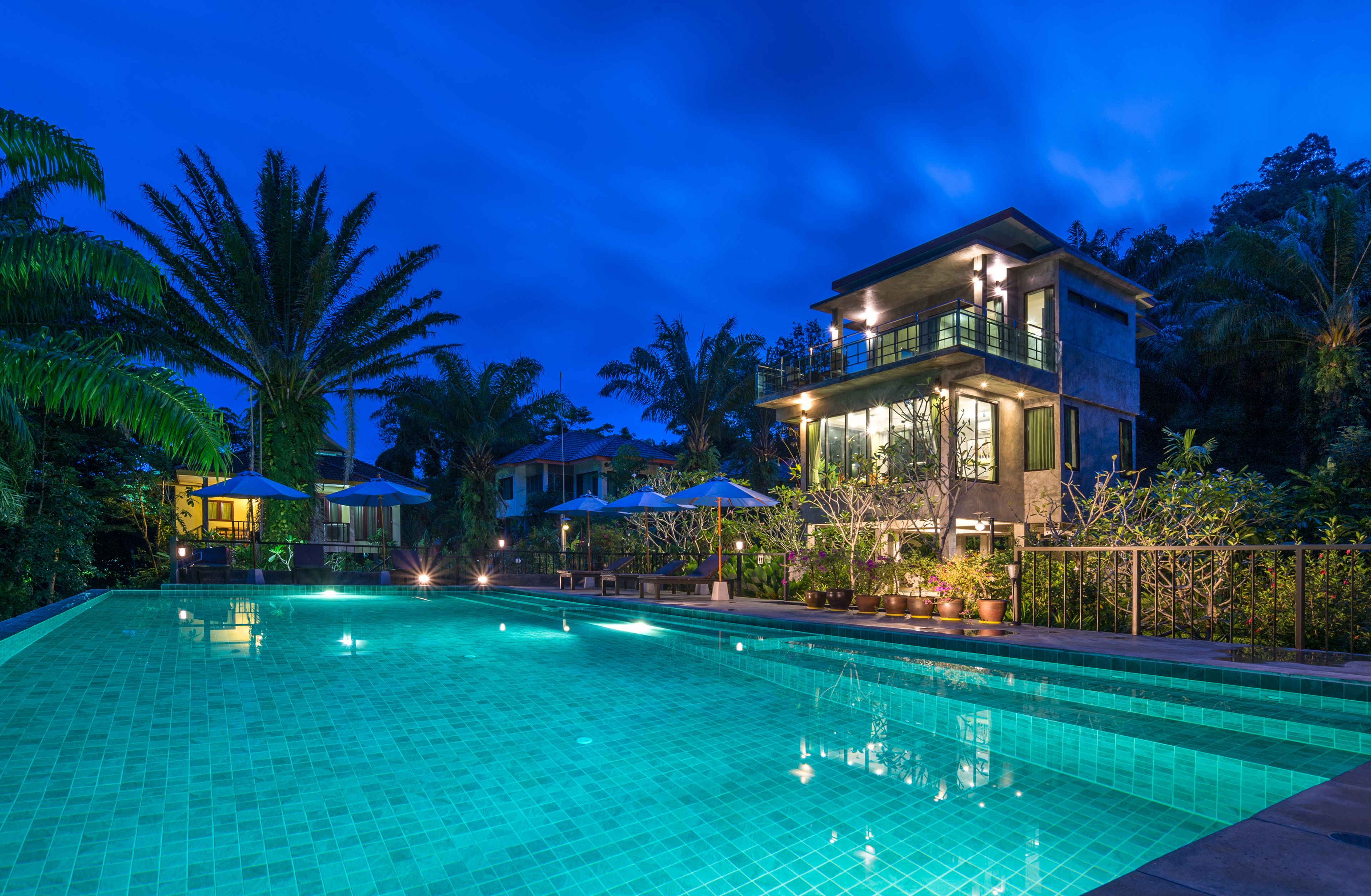 A swimming pool with a tiled surface, surrounded by palm trees and a building with a balcony.