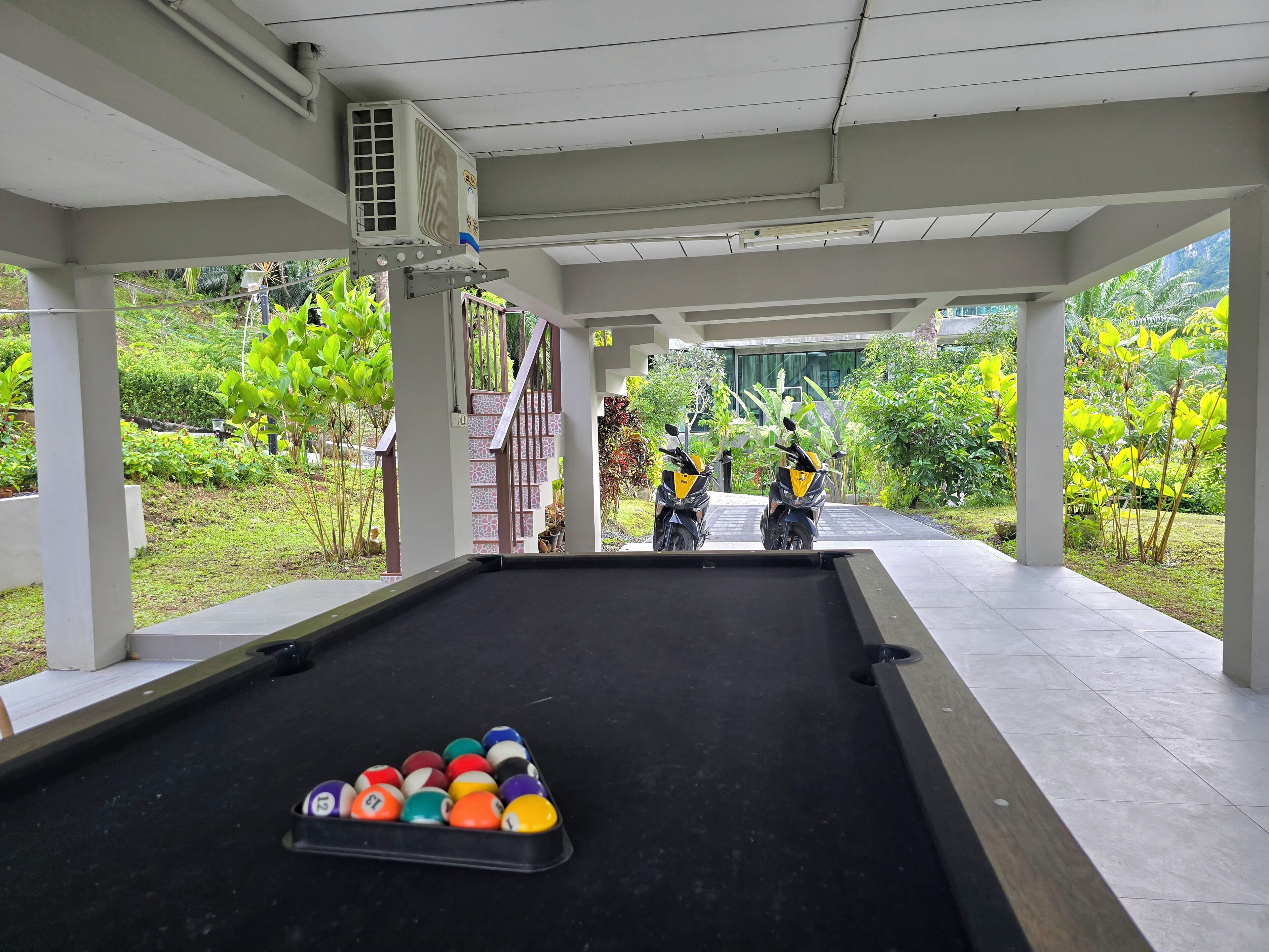 A pool table with billiard balls in a covered area with two motorcycles parked outside.