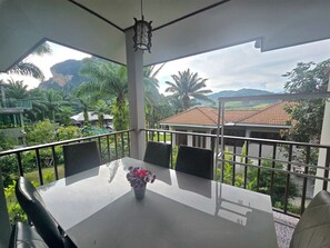 A balcony with a table and chairs, a vase with flowers, and a view of a house and greenery. - Krabi Green Hill Pool Villas (Krabi)