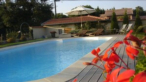 Una piscina con muelle de madera, sillas de descanso y una casa con techo de tejas al fondo.