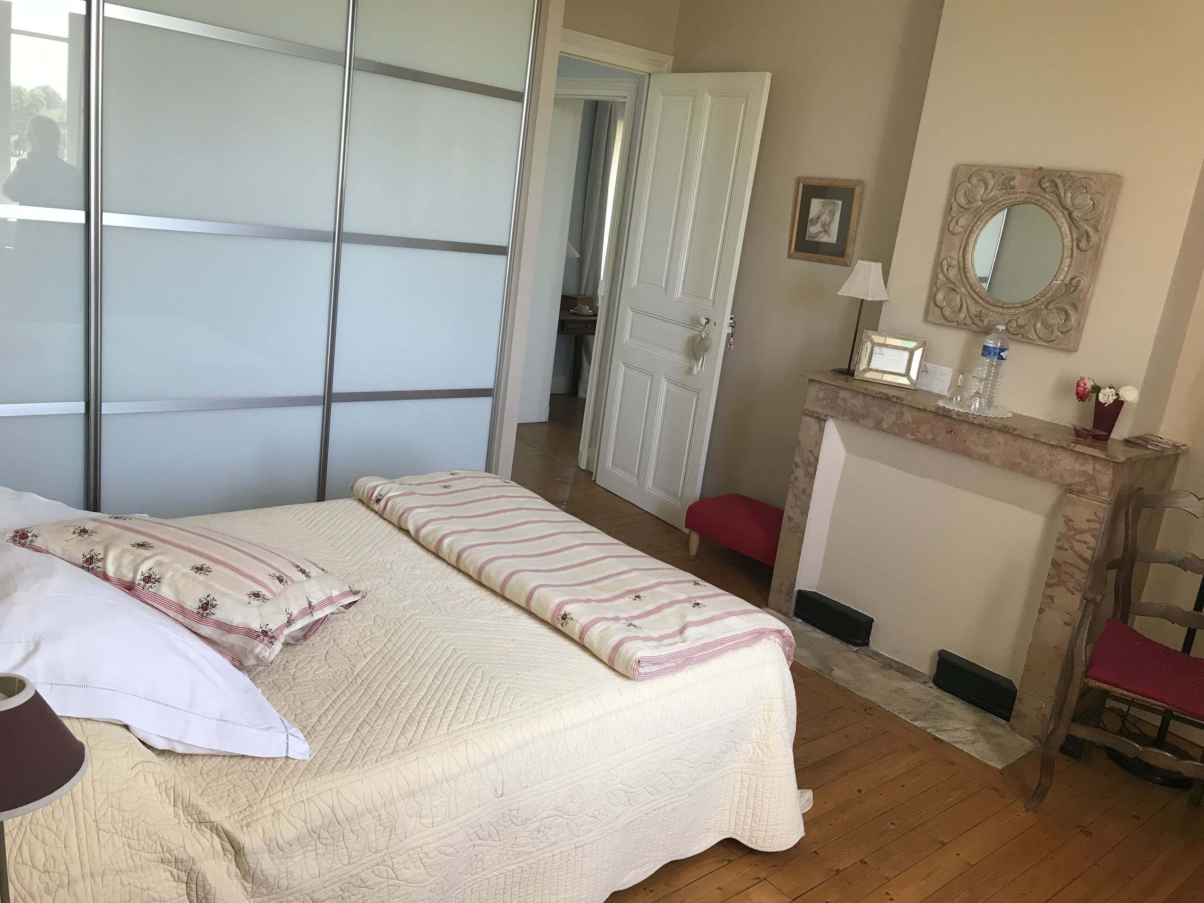 A bedroom with a bed, a bench, a mirror, and a fireplace.