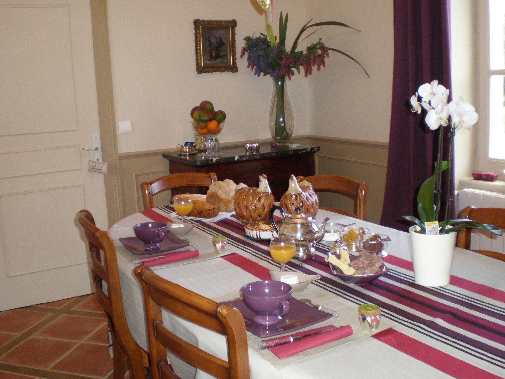 Una mesa de comedor dispuesta con cosas para el desayuno, incluyendo pan, pasteles y un florero con flores.