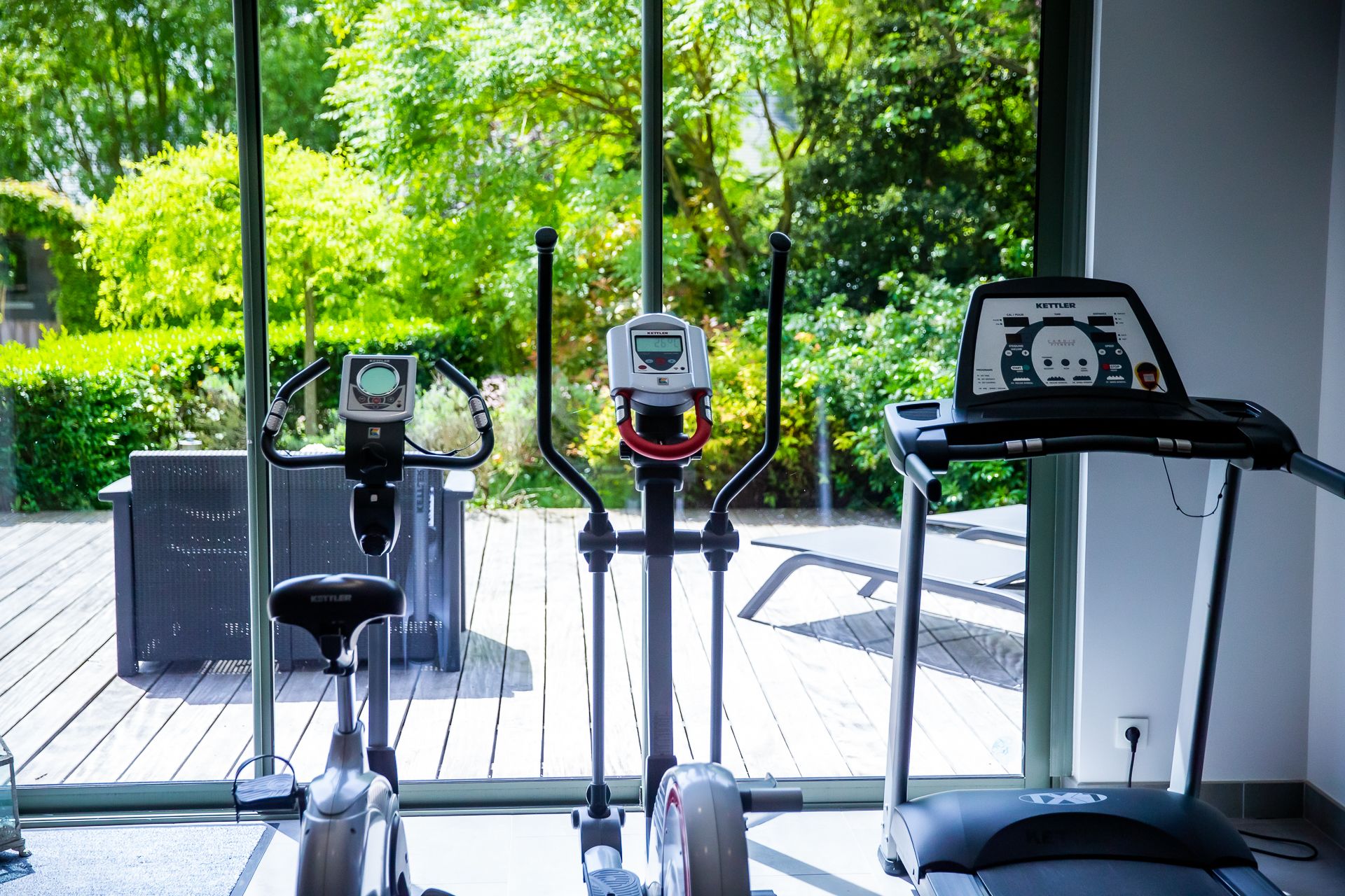 Three stationary bikes with digital displays, positioned in a room with large windows overlooking a wooden deck and lush greenery.