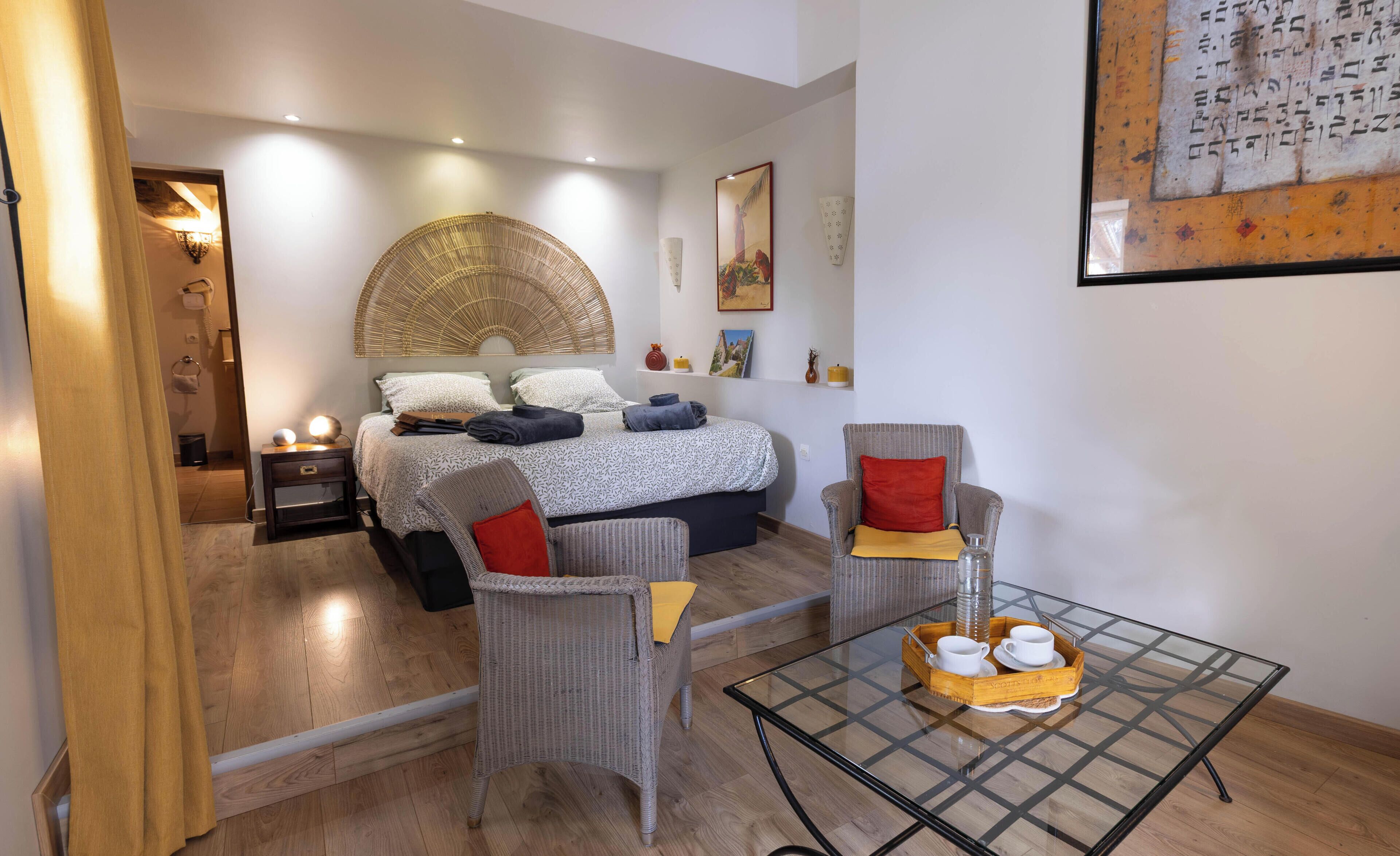 A modern bedroom with a bed, bedside tables, a chair, a glass table with cups, and a wall-mounted artwork.