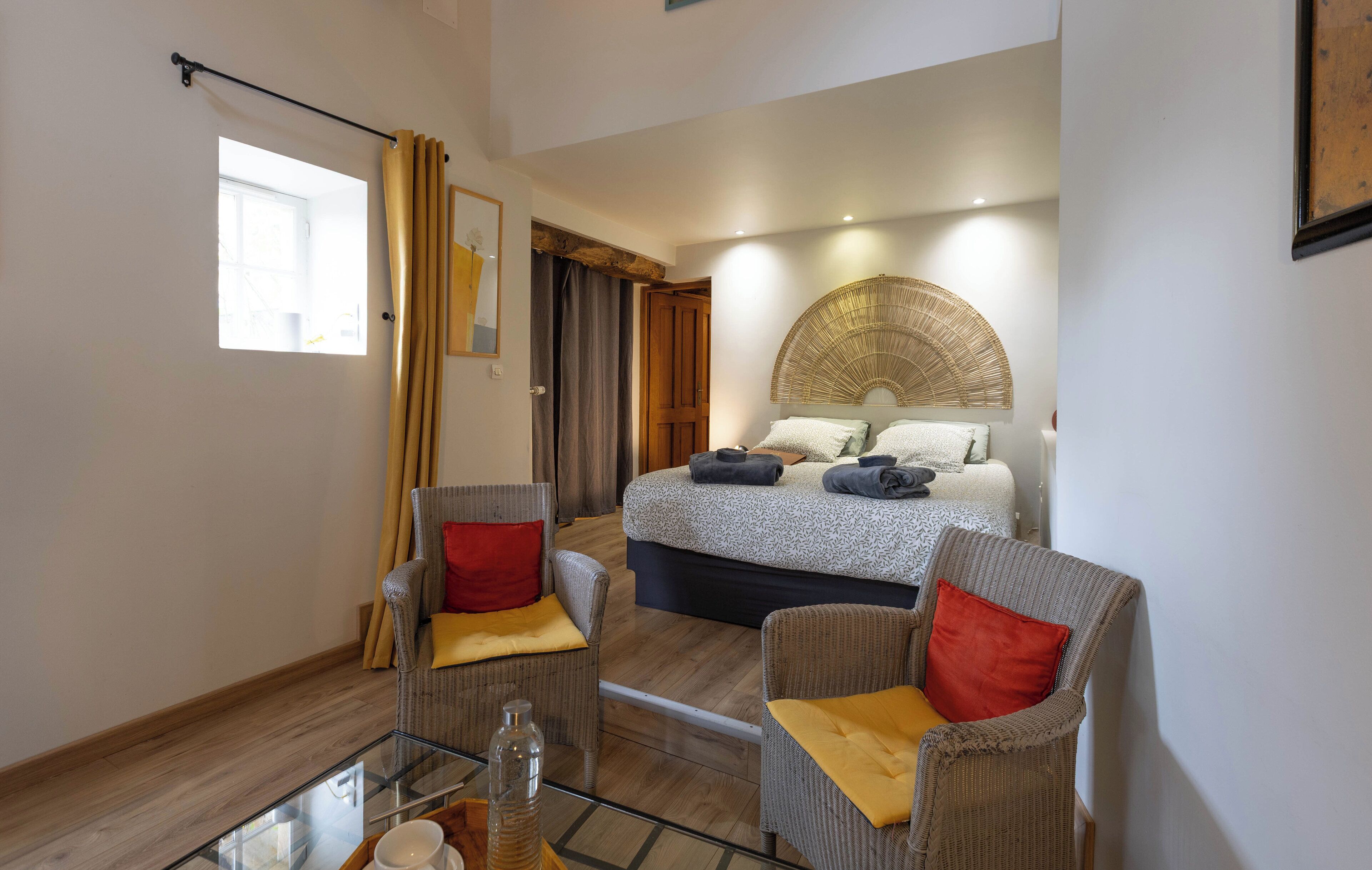 A modern hotel room with a bed, two armchairs, a glass coffee table, and a large window.