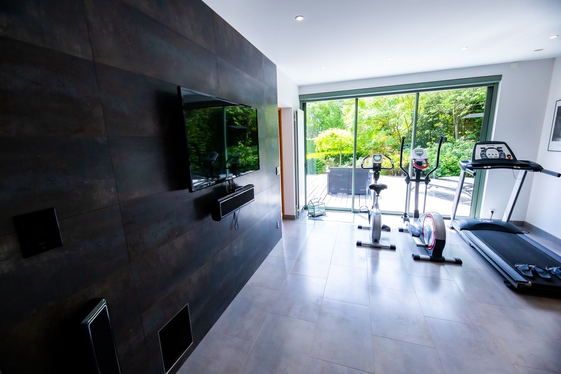 Un gimnasio moderno en casa con un televisor grande, equipo de ejercicio y vistas al exterior con zonas verdes.