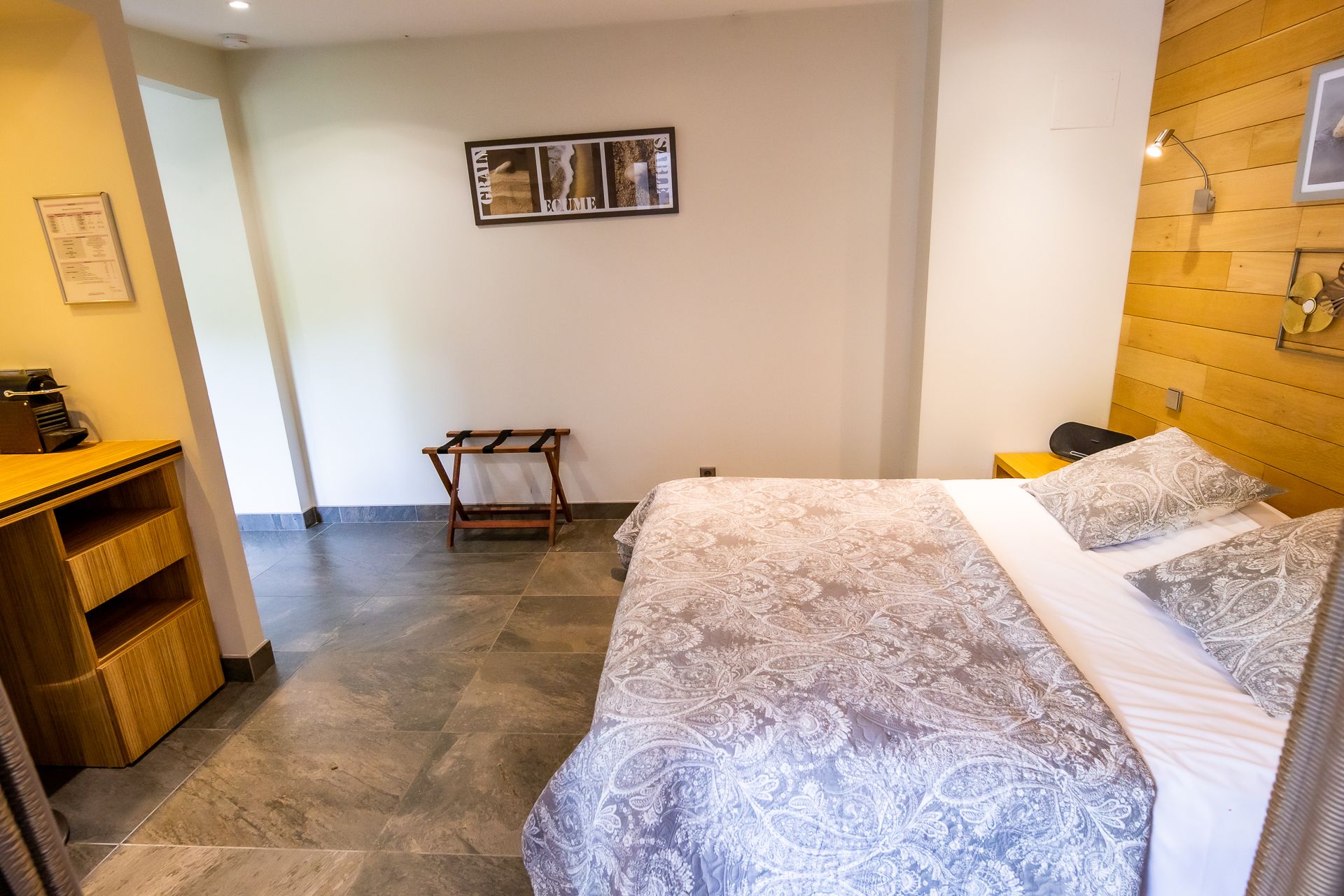 A hotel room with a bed, a wooden headboard, a small table, a chair, a coat rack, and a wall-mounted picture.
