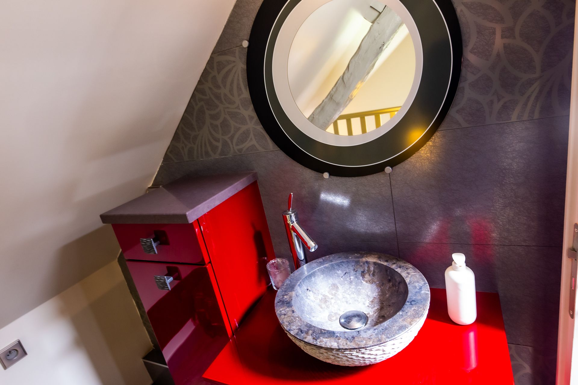 A bathroom with a red vanity, a round mirror, and a stone sink.