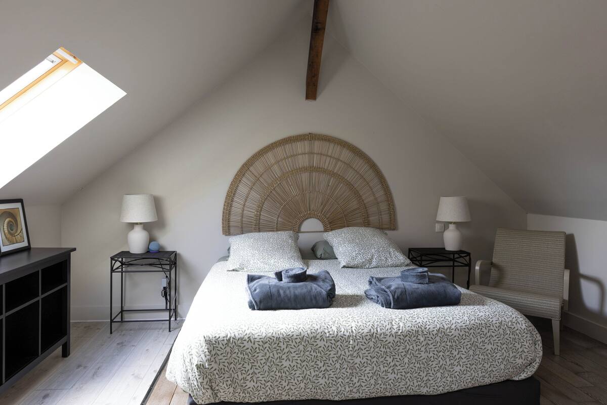 Ein Schlafzimmer mit schrägen Wänden, einem Bett mit gemustertem Überwurf, zwei Nachttischen mit Lampen, einem Sessel und einem kleinen Tisch mit Bilderrahmen.