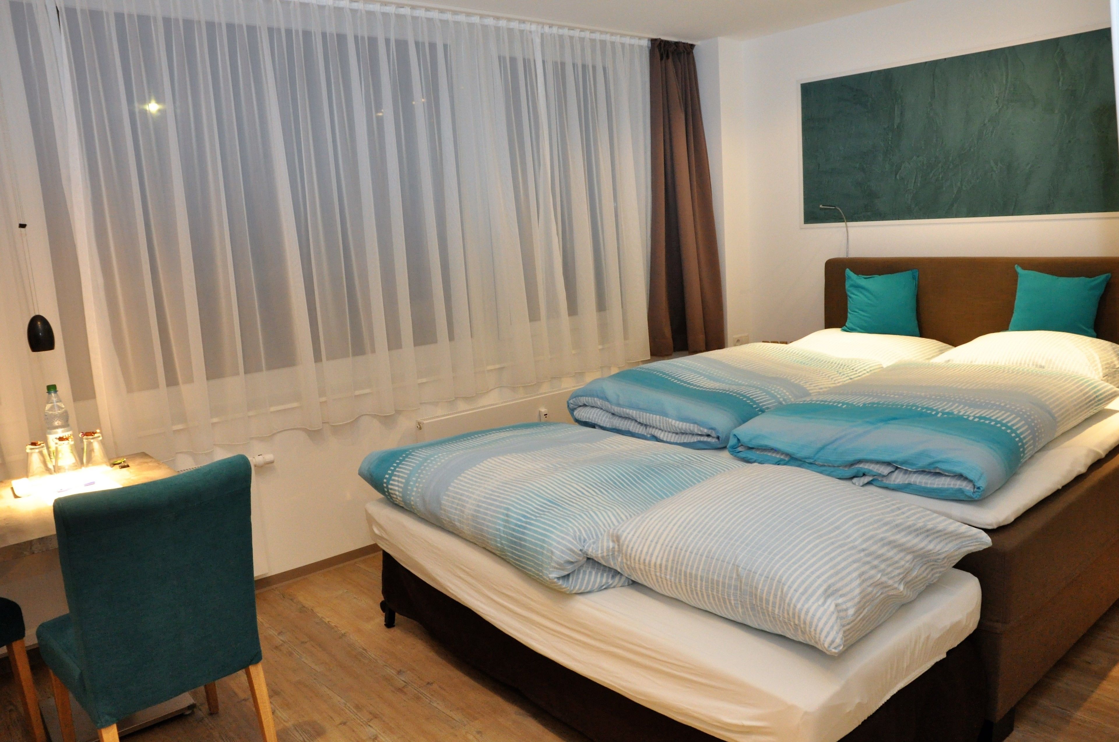 Ein Hotelzimmer mit Bett, Schreibtisch und Stuhl. Ein Fenster mit durchsichtigen Vorhängen und einem teilweise heruntergezogenen braunen Vorhang.