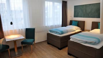 A hotel room with two beds, a small table, a chair, and a window with sheer curtains.