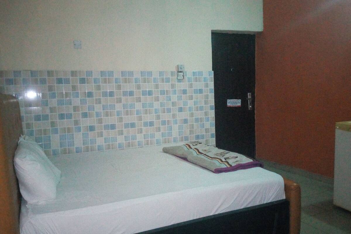 A single bed with a white sheet and a decorative pillow, a nightstand, a small refrigerator, and a tiled wall with a door in the background.