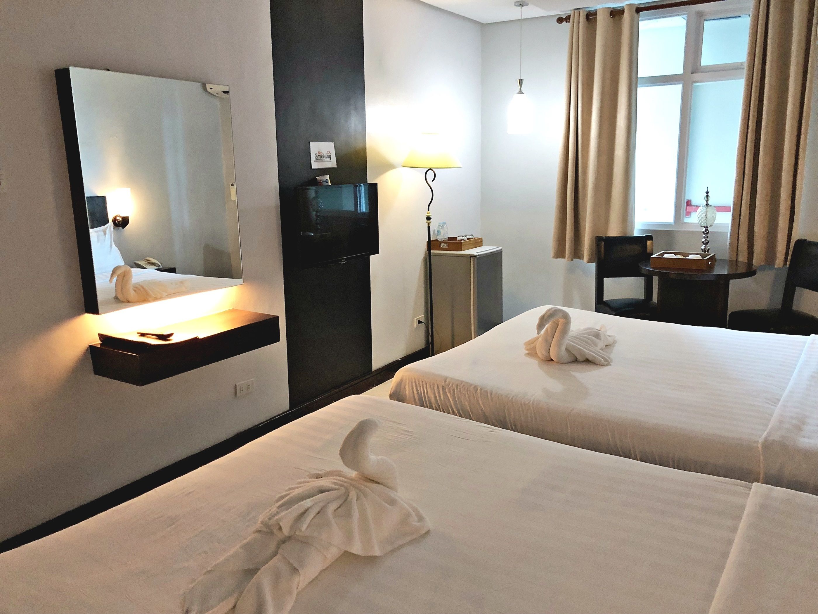 Habitación de hotel con dos camas, un escritorio, una silla, televisor, espejo y ventana con cortinas.