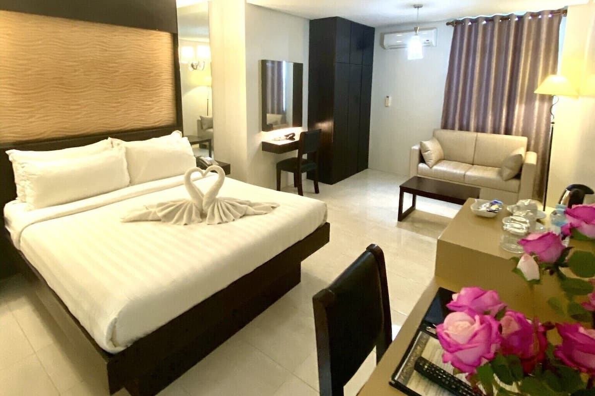 A hotel room with a bed, a desk, a chair, a sofa, and a dining table with flowers.