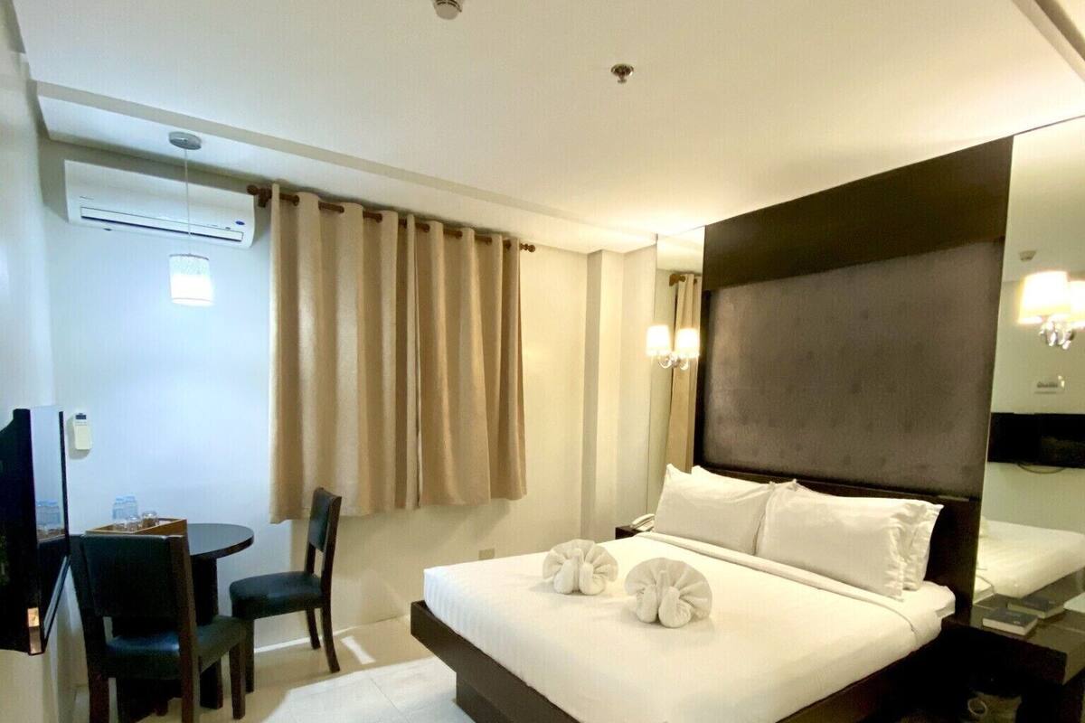 A hotel room with a bed, a desk, chairs, a mirror, and an air conditioning unit.