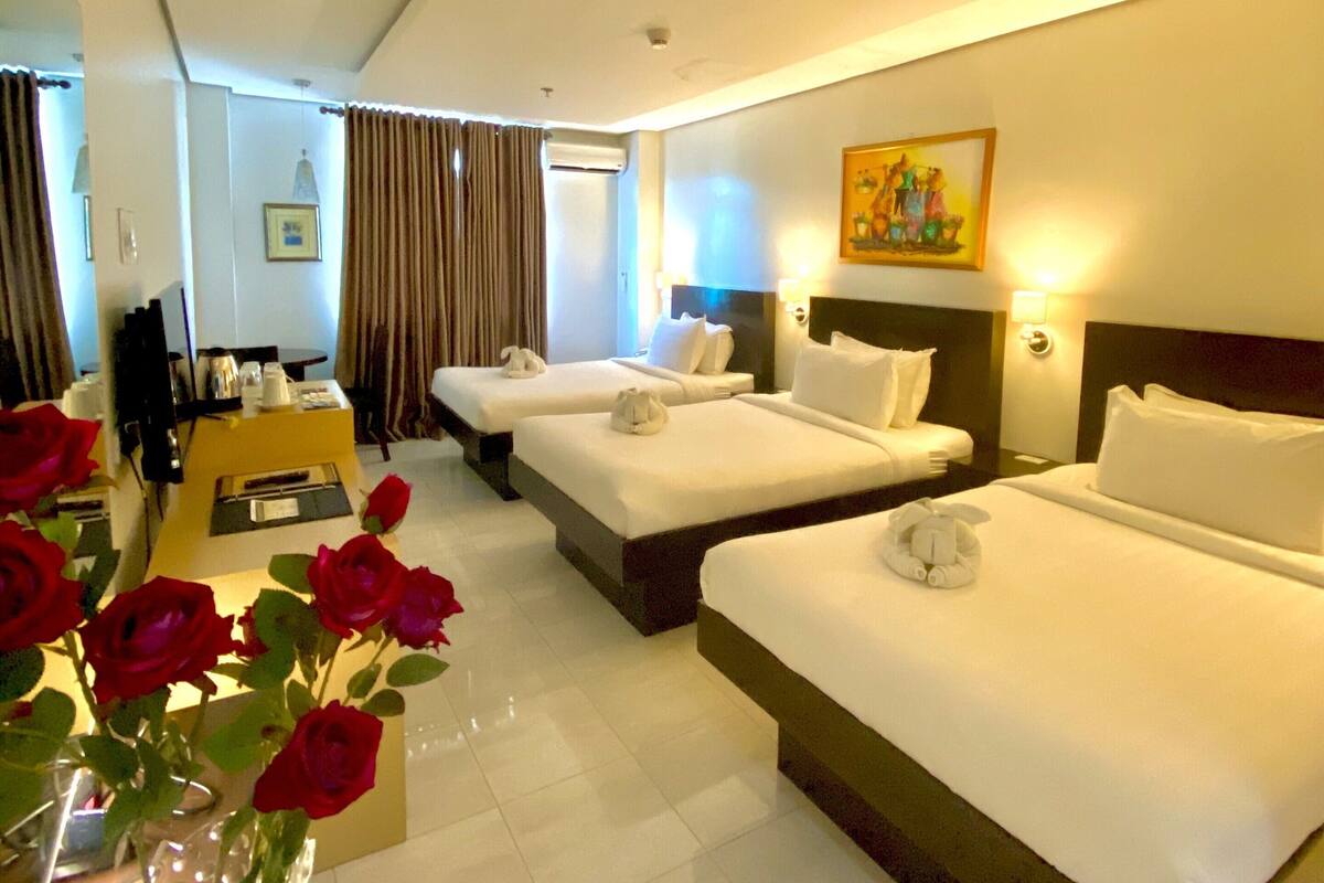 A hotel room with two beds, a desk, a chair, a vase with red roses, and a painting on the wall.