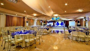 Un salón de banquetes con mesas redondas y sillas, un escenario con un telón de fondo azul y una pantalla grande.
