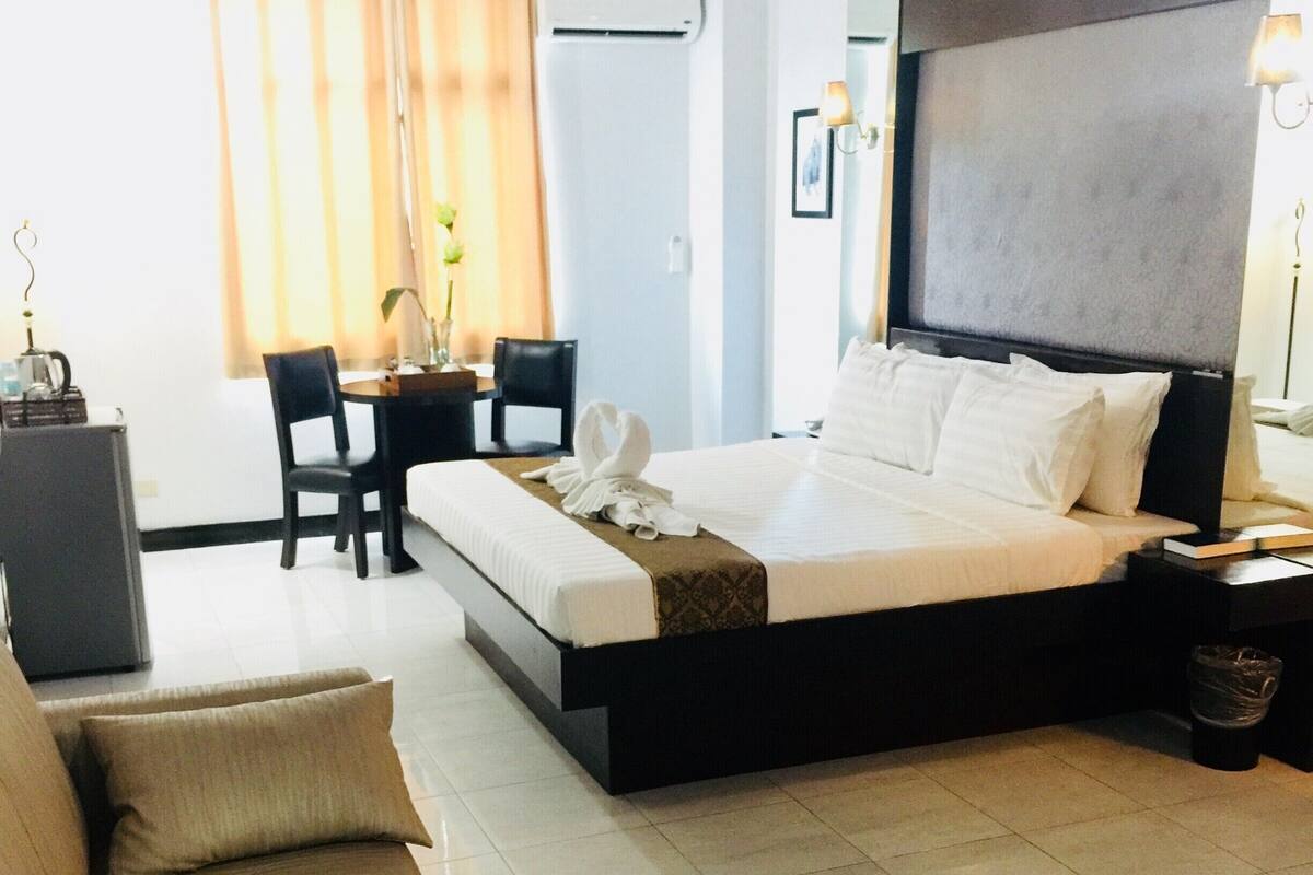 A modern hotel room with a large bed, a sitting area with a sofa and chairs, a dining table, and a TV.