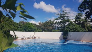 Piscina azul cristalina con sillas de descanso y una escalera, rodeada de vegetación y un muro blanco.