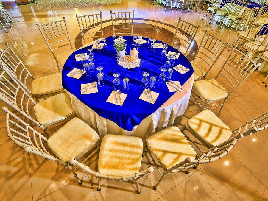 A round table with a blue tablecloth, surrounded by white cushioned chairs with metal frames.