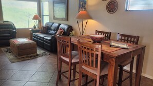 A living room with a black leather sofa, a wooden dining table with chairs, a coffee table, and a rug. There are wall decorations and a window with blinds. - Picture Window Perfect-1 bedrm/den Stunning Mountain View- Many upgrades!!! (Tucson)