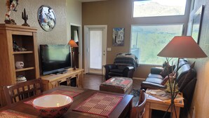 A living room with a TV, wooden furniture, a dining table with placemats, a sofa, and a window with blinds. - Picture Window Perfect-1 bedrm/den Stunning Mountain View- Many upgrades!!! (Tucson)