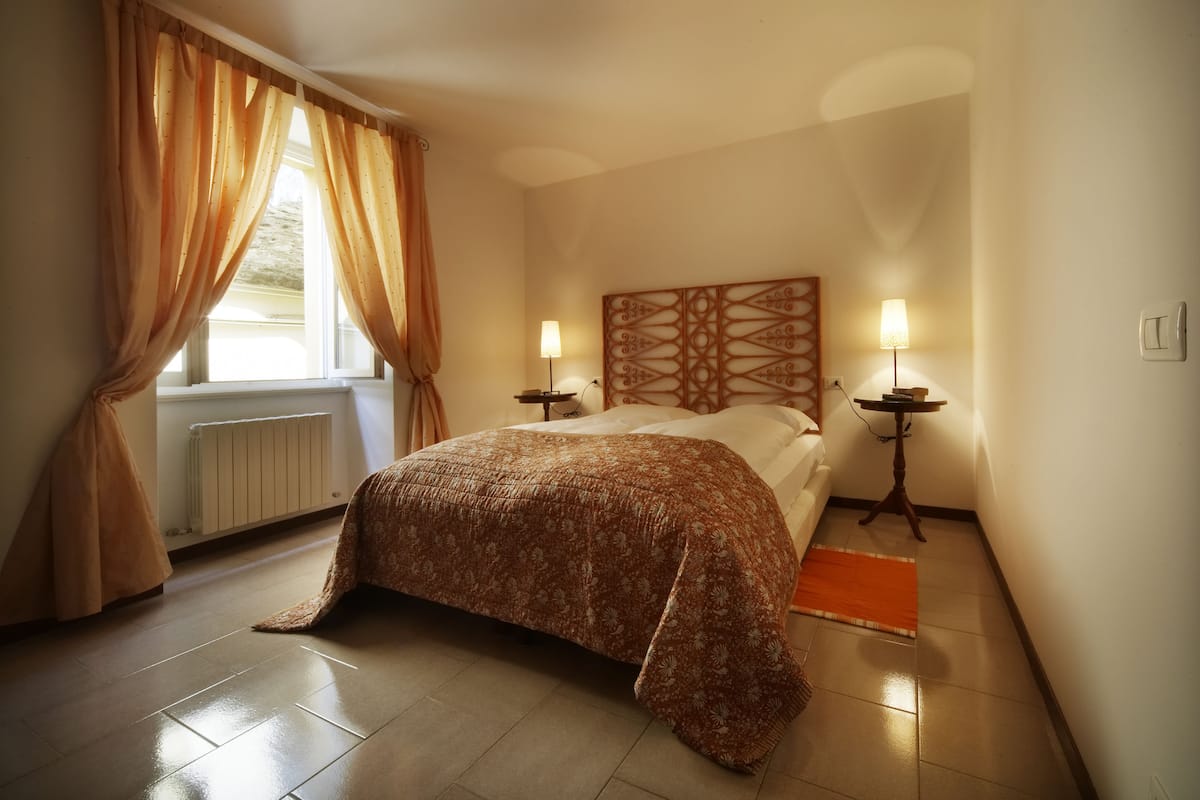Une chambre à coucher avec un lit, deux tables de chevet avec des lampes, un radiateur et une fenêtre avec des rideaux.