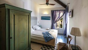 Un dormitorio con una cama, un armario de madera, un ventilador de techo, una lámpara y vistas al exterior a través de una ventana con cortinas.